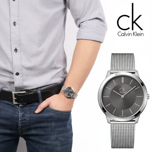 A Calvin Klein watch with a dark gray dial and silver mesh band, shown on a model's wrist alongside a clean product shot of the watch against a white background.